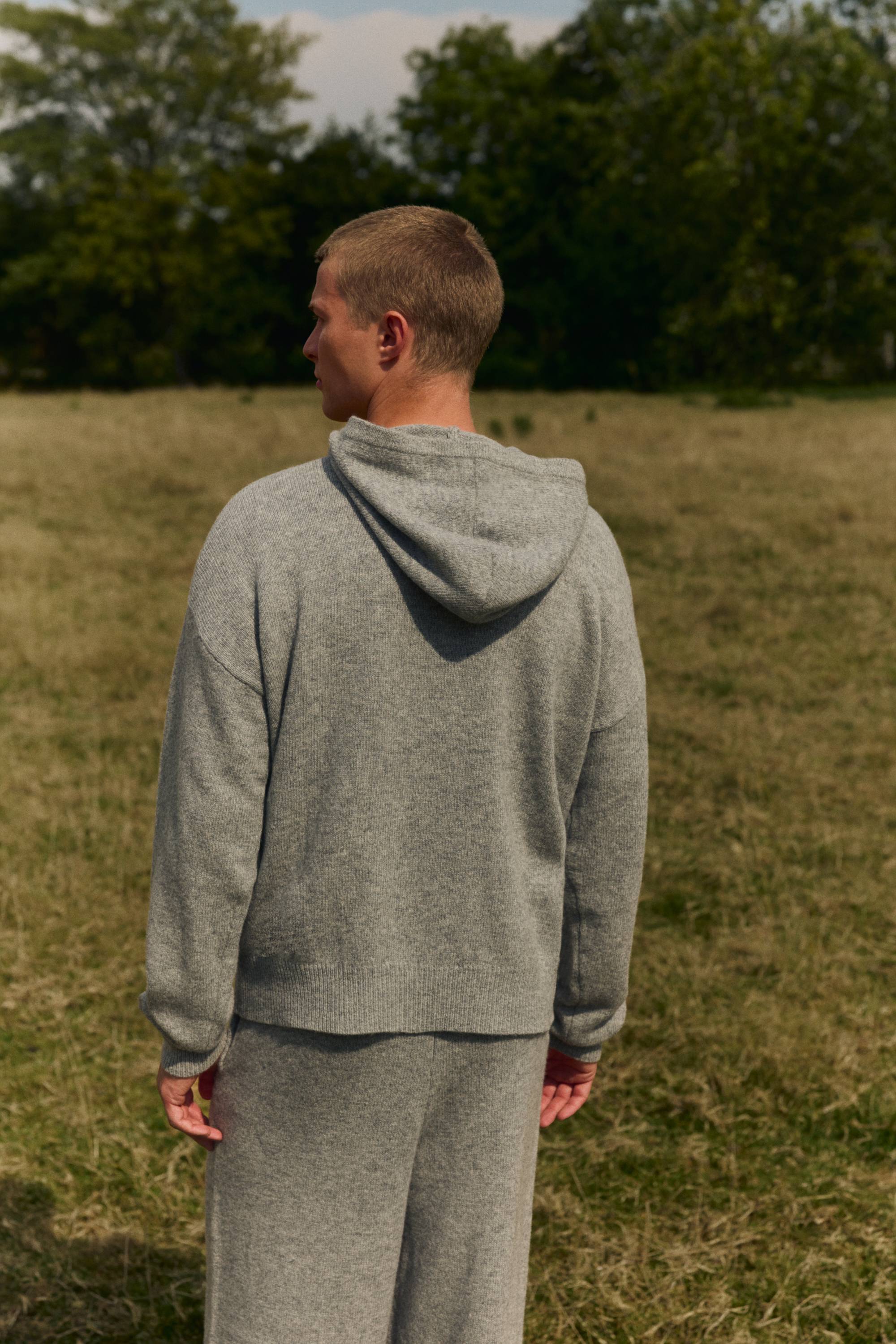 Back view of a man wearing a grey melange CASPARA woolknit hoodie in an open grassy field.
